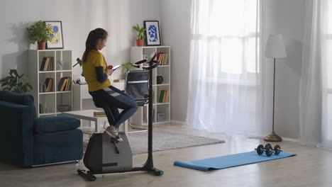 woman-is-typing-and-sending-messages-by-cell-phone-sitting-on-exercise-bike-in-living-room-of-her-flat-break-at-morning-workout-sporty-and-helathy-lifestyle